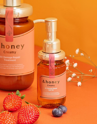 Two bottles of &honey Creamy EX Damage Repair hair oil and treatment, displayed on an orange background with fresh strawberries, blueberries, and delicate white flowers, representing authentic K-beauty skincare available at Sukoshi Mart. 